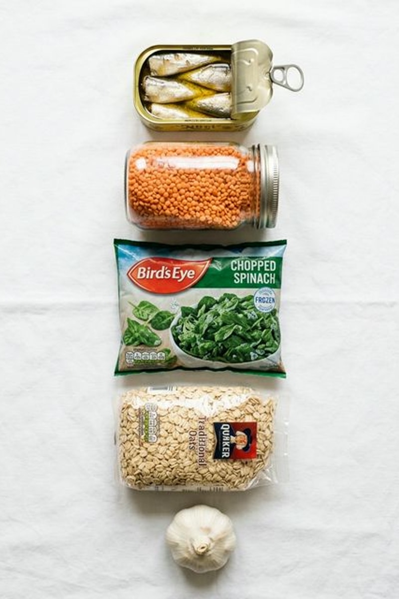 Mediterranean grocery list budget ingredients vertical flat-lay: open sardine tin in olive oil, glass jar of dried red lentils, bag of frozen spinach, rolled oats bag, whole garlic head — clean natural overhead light