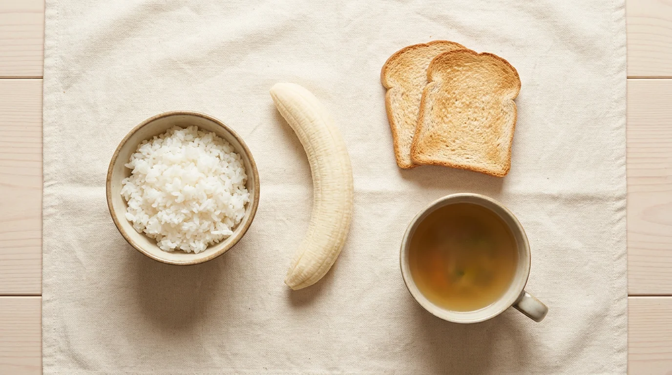Plain recovery foods on cream linen: white rice, a banana, dry toast and a mug of clear broth